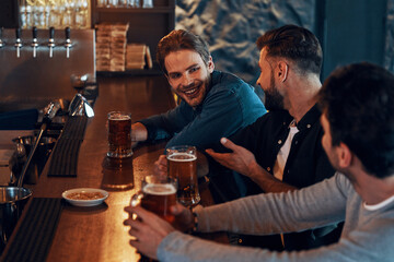 Happy young men in casual clothing drinking beer and bonding together while sitting in the pub