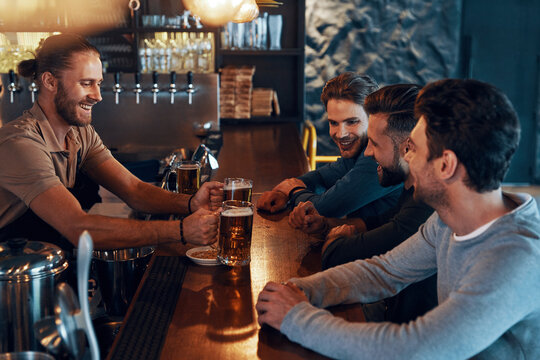 Smiling young men in casual clothing drinking beer and bonding together while sitting in the pub