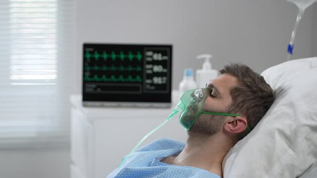 The man lies on the bed in the hospital in oxygen masks and regains consciousness. Connected to the breathing and heart rate monitor lies in the hospital on the bed with his eyes closed