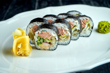 Appetizing Japanese futomak sushi roll with salmon, cucumber in a white plate on a black wooden background with ginger and wasabi. Japanese cuisine. Futomak roll