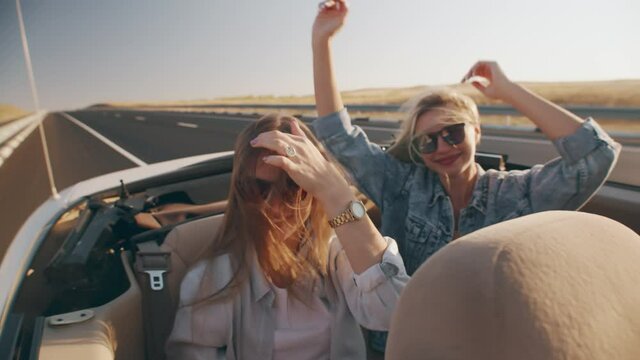 Two beautiful young women ride in white convertible driving on highway, against background of field. Women enjoy travel and freedom