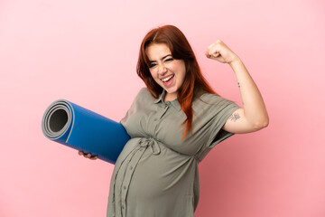 Young redhead caucasian woman isolated on pink background pregnant and doing strong gesture while going to yoga classes