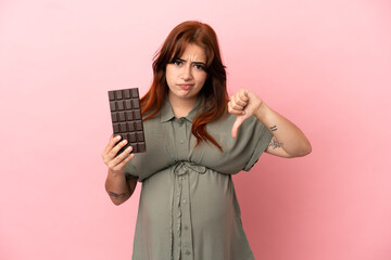 Young redhead caucasian woman isolated on pink background pregnant and holding chocolate doing bad signal