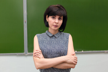 A brunette schoolteacher stands at the blackboard with her arms folded.