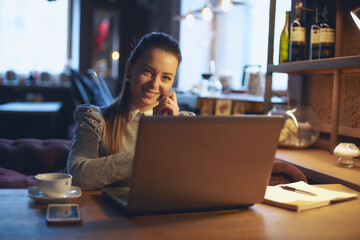 Young beautiful woman sits at a wooden table in a cafe indoors and works on a laptop. Conception of remote work and freelance work.