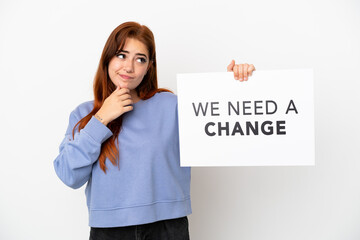 Young redhead woman isolated on white background holding a placard with text We Need a Change and thinking