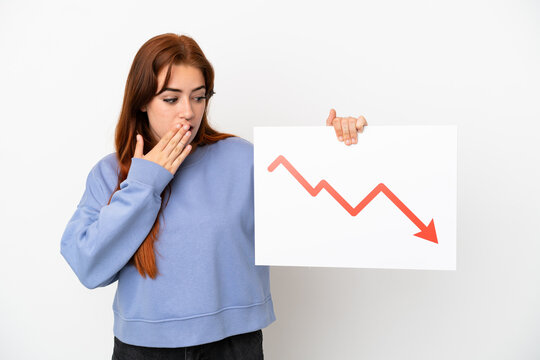 Young redhead woman isolated on white background holding a sign with a decreasing statistics arrow symbol with surprised expression