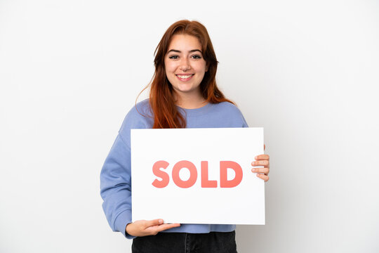 Young redhead woman isolated on white background holding a placard with text SOLD with happy expression