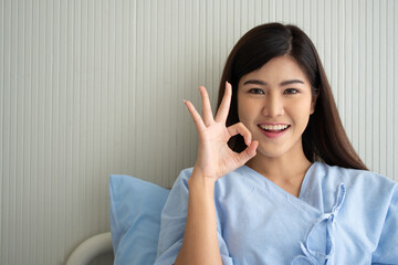 Happy Asian female patient smiling, lies on the bed, and Shows an ok symbol to show confidence in treatment. Concept of believe in treatment And insurance coverage