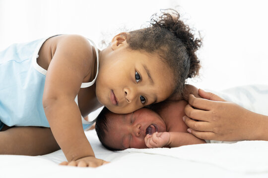 African American little girl playing and comforting newborn baby on white bed. Little girl takes care of infant baby with kindly