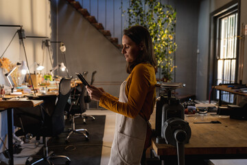 Caucasian female jeweller wearing apron, standing in workshop, using tablet, smiling