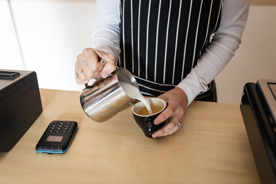 Midsection of caucasian female business owner wearing apron, pouring froth milk to coffee