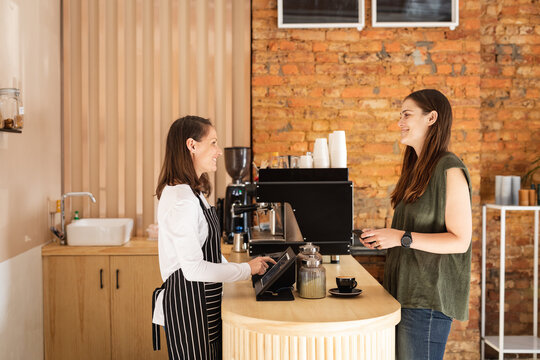 Two caucasian female business owner and customer standing at countertop, talking and smiling