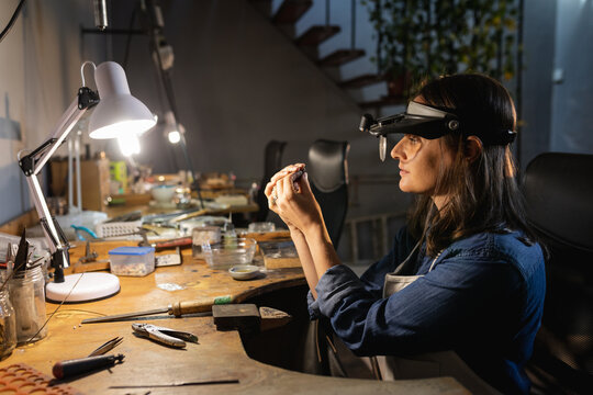 Caucasian female jeweller sitting at desk, holding jewelry tools, making jewelry in workshop