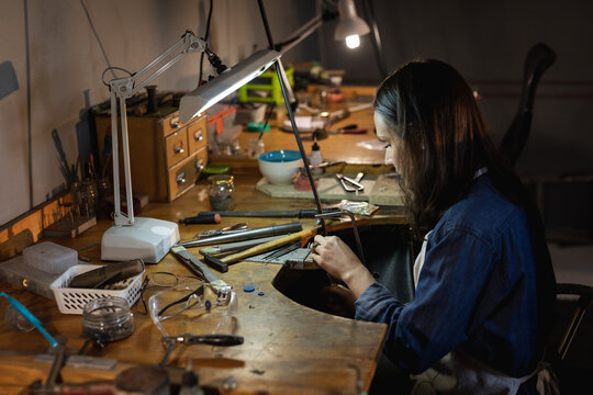 Caucasian female jeweller sitting at desk, holding jewelry tools, making jewelry in workshop