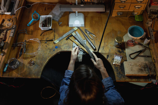 Caucasian female jeweller sitting at desk, holding jewelry tools, making jewelry in workshop
