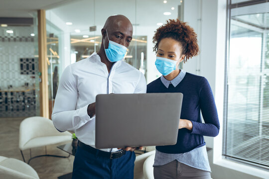 Two diverse business colleagues wearing face masks and using laptop