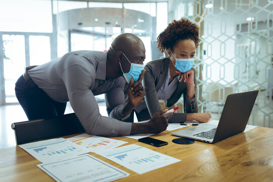Two diverse male and female business colleagues wearing face masks and using laptop