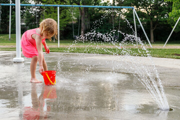 Little girl playing at water splash pad fountain in the park playground during hot summer day.