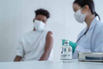 Selective focus on Covid-19 vaccine bottle on table. Blur background is Asian female doctor with mask and gloves is putting plaster on African American boy arm after vaccinated.