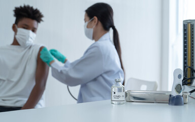Fototapeta premium Selective focus on Covid-19 vaccine bottle on table. And next to it is thermometer and blood pressure monitor. Blur background is Asian woman doctor putting plaster on African boy arm after vaccinated