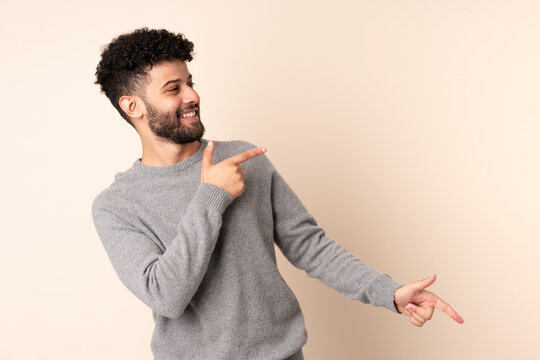 Young Moroccan man isolated on beige background pointing finger to the side and presenting a product
