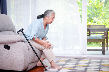 Asian elderly woman Sitting on the sofa in the house, there was a walking stick nearby. and had knee pain, unable to walk. concept of elderly health problems Prevention and health care in retirement.