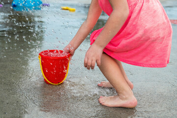 Little girl playing barefoot at water splash pad fountain in the park playground on a hot summer day.