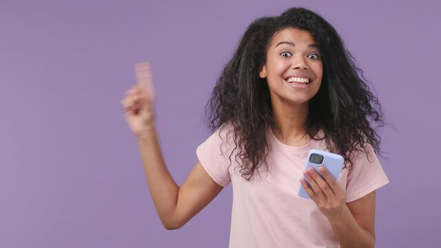 Young african woman 20s years old in pink T-shirt hold use mobile cell phone point finger hand aside on workspace copy space mockup promo area isolated on pastel violet purple color background studio