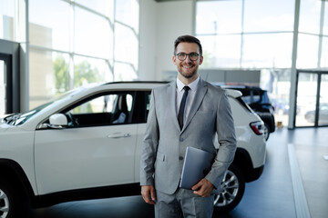 Confident young car dealer standing in showroom and holding laptop computer while posing. - obrazy, fototapety, plakaty