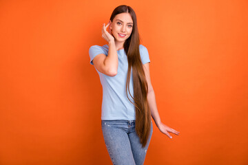 Photo of cute millennial lady touch hair wear blue t-shirt jeans isolated on orange color background