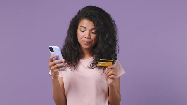 Young african woman in pink T-shirt use mobile cell phone hold in hand credit bank card doing online shopping order delivery to home book tour isolated on pastel violet purple color background studio