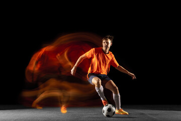 Fototapeta na wymiar Young caucasian male football or soccer player kicking ball for the goal in mixed light on dark background. Concept of healthy lifestyle, professional sport, hobby.