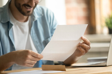 Close up young man holding paper correspondence in hands, reading letter with good news, getting...