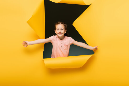 Joyful Kid With Outstretched Hands Looking At Camera Through Ripped Hole On Yellow Background.