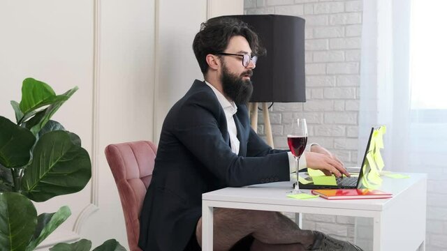 Bearded man with no pants works at the computer, the man types then close the computer and enjoy a glass of wine and looks happily. Enjoy success after a hard day's work.