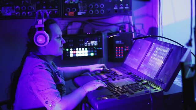 A woman lighting engineer in headphones controls lighting devices on stage at the remote control in the lighting room.