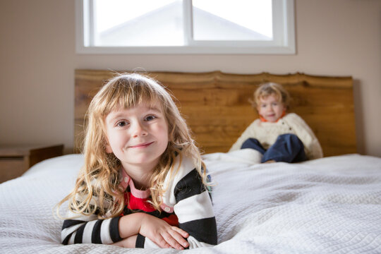 Girl lays on bed smiling for camera with brother sitting behind her