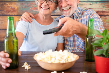 Happy retired couple watching at football match sitting at pub with two bottles of beer and some popcorn. Two attractive senior people