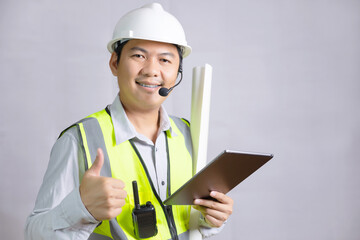 A young man, architect, or engineer wearing a reflector and a safety helmet for construction work, stands holding a design blueprint with a tablet and looking at the camera.