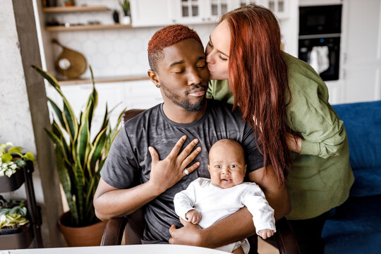 Happy african American American young father sit with small boy son child, spend time with young caucasian wife, kissing husband, mixed race family with child at home , smile enjoy weekend together