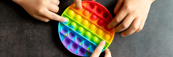 anti stress sensory pop it toys in a children's hands. a little happy kids plays with a simple dimple toy on a black table. toddlers holding and playing popit rainbow color, trend 2021 year. banner