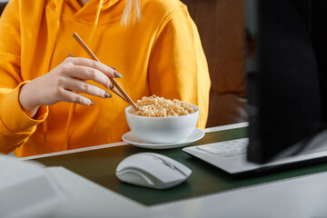 Woman eats noodles with chopsticks behind computer at home workplace, home office during video game...