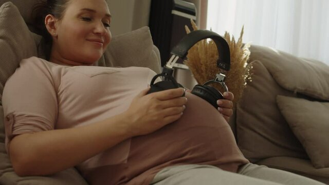 Adult pregnant woman listens to music with headphones. Happy pregnant woman expecting a baby. Pregnant woman feeling happy at home while. Woman pregnancy concept and maternity prenatal care. 4K.