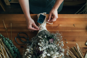 Hands of Female Florist Making Everlasting Bouquet of Dried Flowers at Wooden Table at Flower...