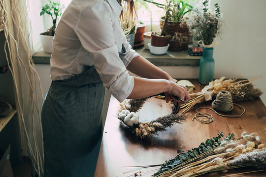 Hands of Female Florist Making Wreath with Dry Flowers at Wooden Table at her Flower Shop.
Side view of unrecognizable woman entrepreneur making decorative wreath with dry flowers and plants.
