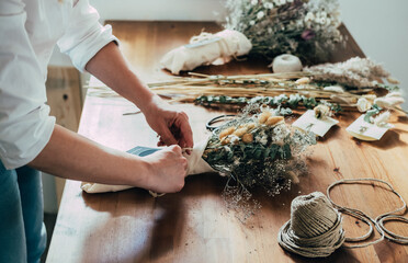Hands of Female Florist Making Everlasting Bouquet of Dried Flowers at Wooden Table at her Flower Shop.
Side view of unrecognizable woman entrepreneur arranging bouquet of a dry flowers and plants. - obrazy, fototapety, plakaty