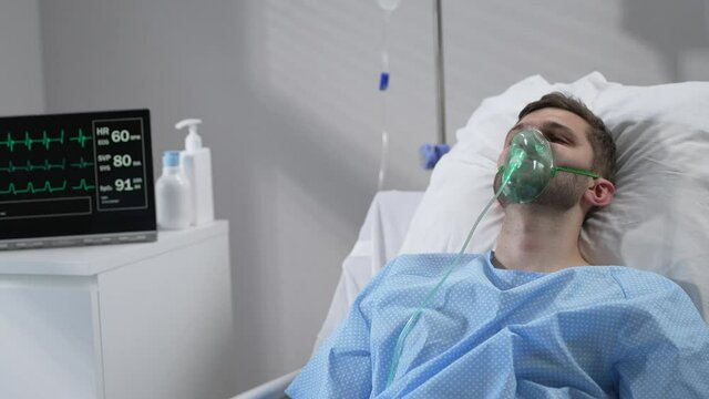 The man lies on the bed in the hospital in oxygen masks and regains consciousness. Connected to the breathing and heart rate monitor lies in the hospital on the bed with his eyes closed