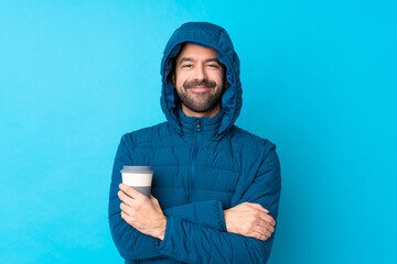 Man wearing winter jacket and holding a takeaway coffee over isolated blue background keeping the arms crossed in frontal position