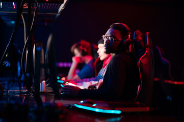 Fototapeta na wymiar A member of the esports team at a bootcamp training session. Portrait of a black guy with glasses at a gaming computer with a glowing keyboard.
